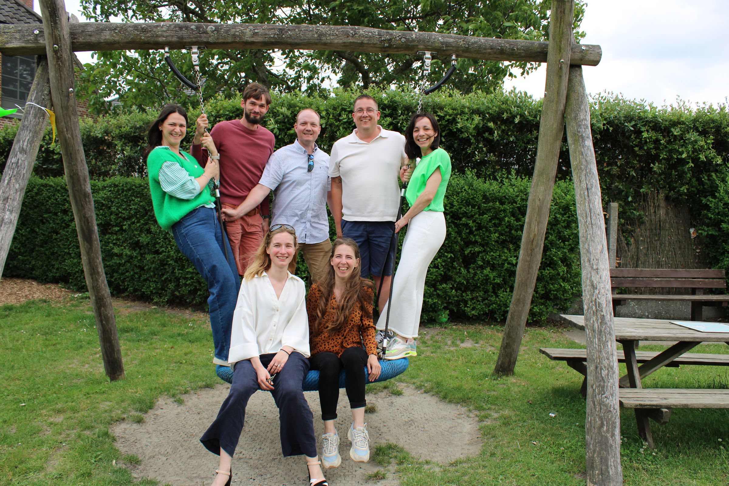 Zes volwassenen zitten en staan op een grote schommel in een tuin tijdens de Care4CMV-familiedag